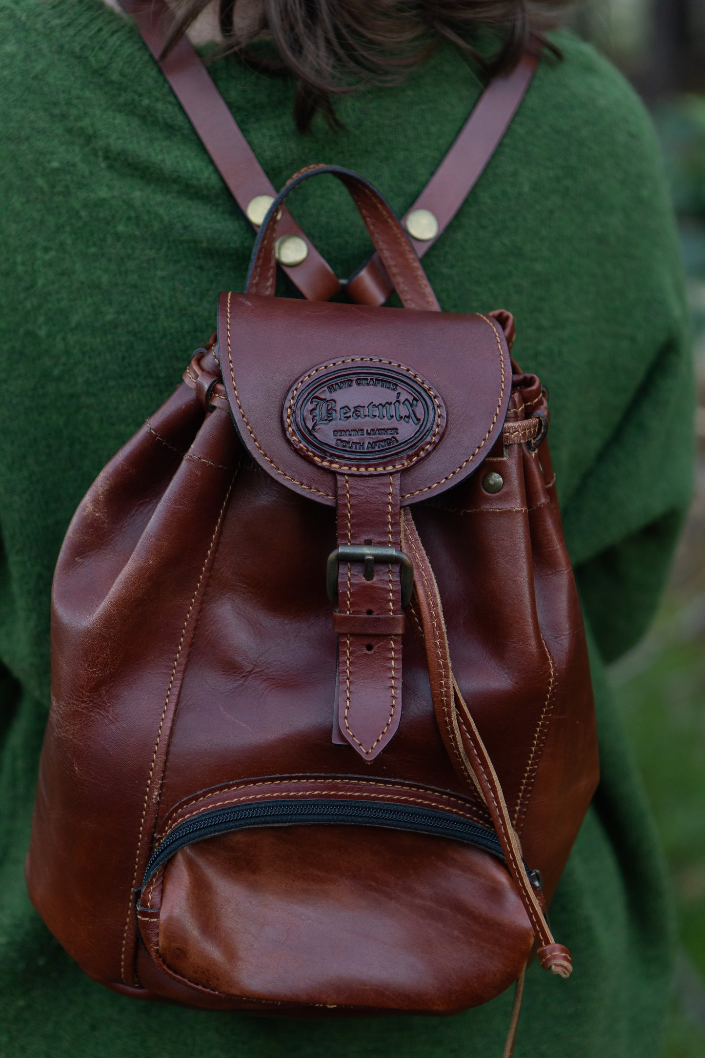 A person wearing a green sweater is carrying a brown leather daypack with a main compartment and a front pocket. The daypack features a leather handle and shoulder straps.