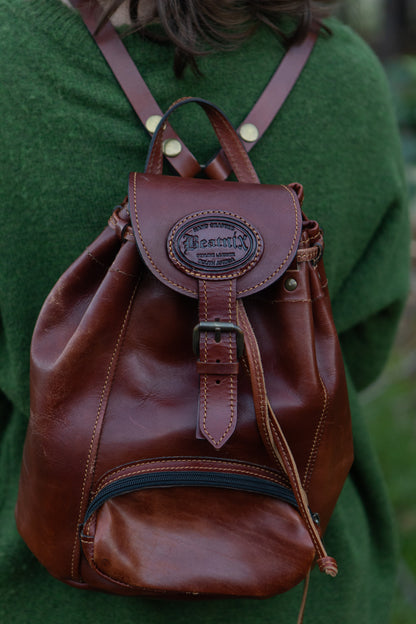 A person wearing a green sweater is carrying a brown leather daypack with a main compartment and a front pocket. The daypack features a leather handle and shoulder straps.