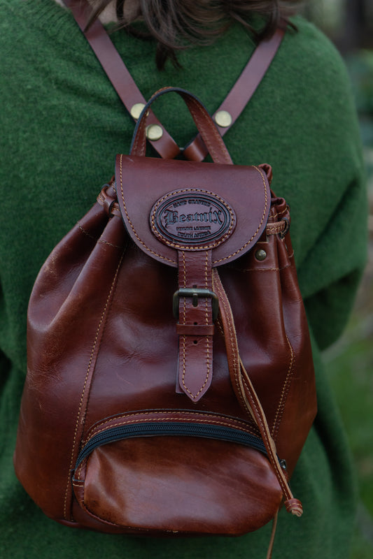 A person wearing a green sweater is carrying a brown leather daypack with a main compartment and a front pocket. The daypack features a leather handle and shoulder straps.